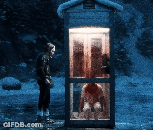 A Man Stands In Front Of A Phone Booth With A Woman Sitting Inside GIF