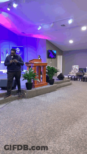 A Man Stands In Front Of A Podium In A Church With Potted Plants GIF