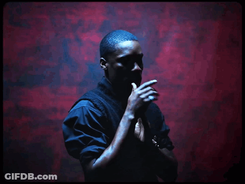 A Man Stands In Front Of A Red Wall With His Hands Folded GIF