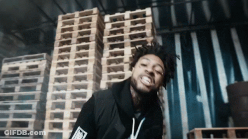 A Man Stands In Front Of A Stack Of Wooden Pallets With The Letter T On His Shirt GIF