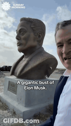 A Man Stands In Front Of A Statue Of Elon Musk And Says A Gigantic Bust Of Elon Musk GIF