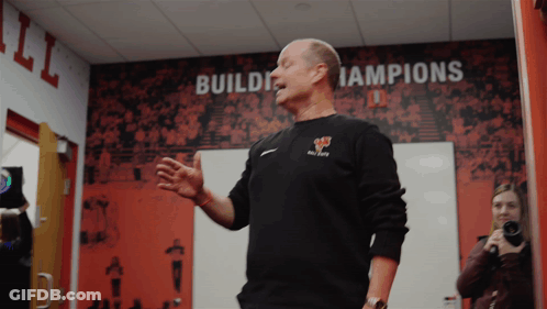 A Man Stands In Front Of A Wall With The Words Build Champions On It GIF
