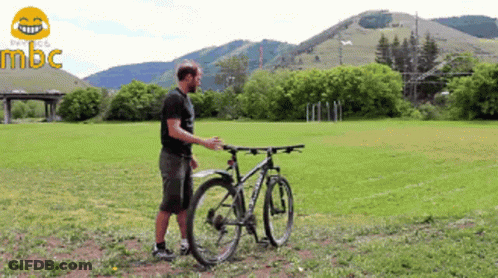 A Man Stands Next To A Bicycle In A Grassy Field With Mountains In The Background And The Word Mbc In The Corner GIF
