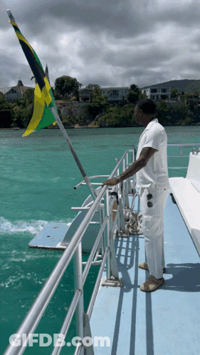 A Man Stands On A Boat With A Flag Flying In The Background GIF
