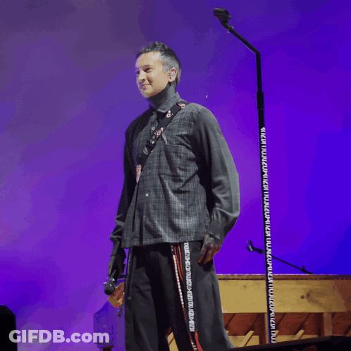 A Man Stands On A Stage Holding A Microphone With A Purple Backdrop GIF