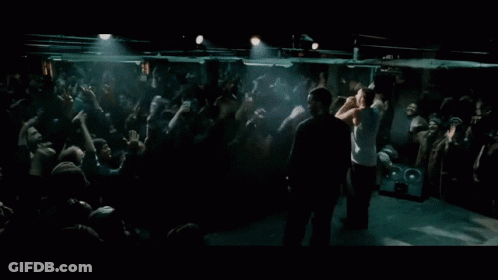 A Man Stands On A Stage In Front Of A Crowd With His Hands In The Air GIF