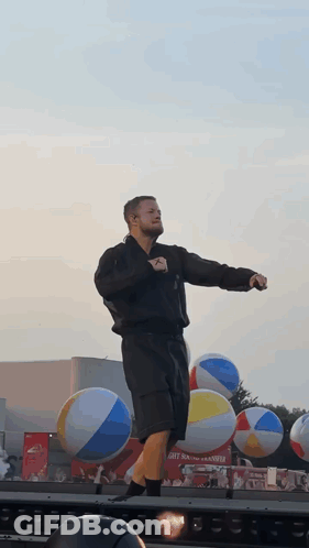 A Man Stands On A Stage In Front Of Beach Balls GIF