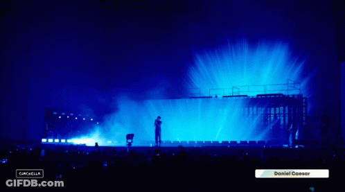 A Man Stands On A Stage With A Blue Light Behind Him And A Sign That Says Daniel Caesar GIF
