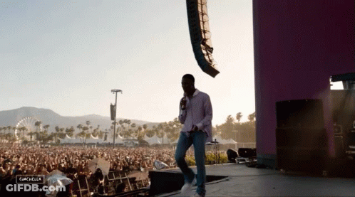 A Man Stands On A Stage With A Microphone In Front Of A Crowd At A Music Festival And The Word Coachella Is Visible GIF
