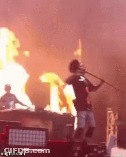 A Man Stands On A Stage With A Microphone In Front Of A Fire GIF
