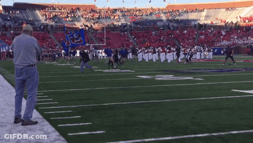 A Man Stands On The Sideline Of A Football Field Watching A Marching Band GIF