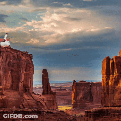 A Man Stands On Top Of A Rocky Cliff Overlooking A Canyon GIF