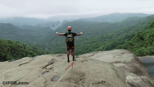 A Man Stands On Top Of A Rocky Cliff With His Arms Outstretched In Front Of A Lush Green Forest .. GIF