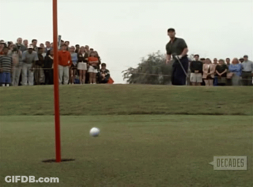 A Man Swings A Golf Club At A Golf Ball On A Golf Course With People Watching GIF