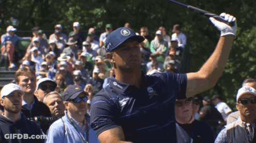 A Man Swings A Golf Club In Front Of A Crowd That Includes A Man Wearing A Hat That Says R On It GIF