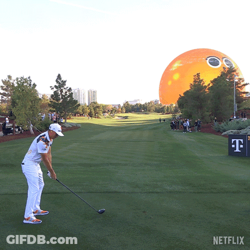A Man Swings A Golf Club On A Golf Course With An Orange Balloon In The Background GIF