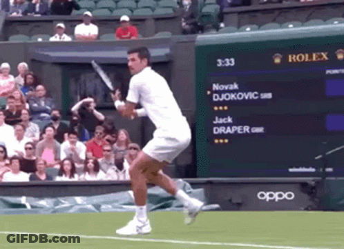 A Man Swings A Tennis Racquet In Front Of A Scoreboard That Says Novak Djokovic GIF