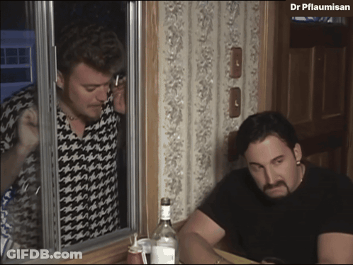 A Man Talking On A Cell Phone Next To A Man Sitting At A Table With A Bottle Of Vodka GIF
