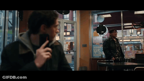 A Man Talking On A Cell Phone While Another Man Stands In Front Of A Sign That Says ' Pepsi ' On It GIF