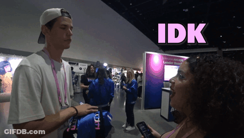 A Man Talking To A Woman In Front Of A Sign That Says Idk On It GIF