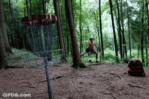 A Man Throwing A Frisbee In The Woods Near A Disc Golf Basket GIF
