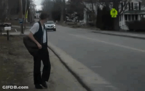 A Man Walking Down A Street With A Car Behind Him And A Yellow Sign That Says Right Turn GIF