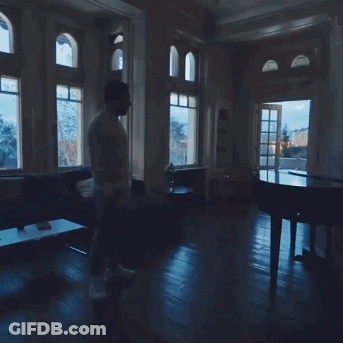 A Man Walking In A Dark Room With A Piano In The Background GIF