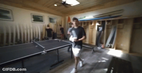 A Man Walking In A Room With A Ping Pong Table In The Background GIF