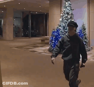 A Man Walking In Front Of A Building With Christmas Trees GIF
