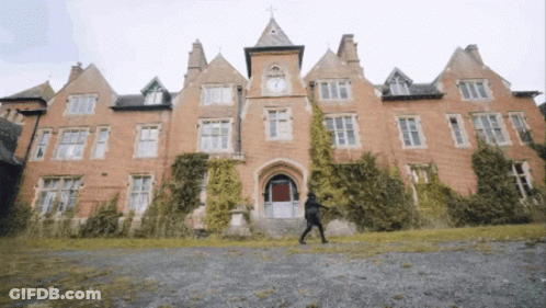 A Man Walking In Front Of A Large Brick Building With A Clock Tower GIF