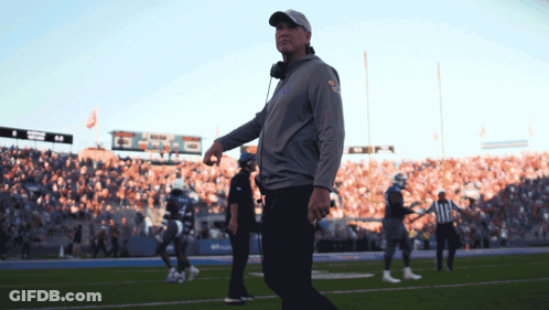 A Man Walking On A Football Field With A Scoreboard In The Background That Says ' Denver ' On It GIF