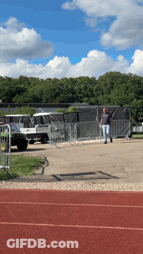 A Man Walking On A Track In Front Of A Fence With A Jeep In The Background GIF