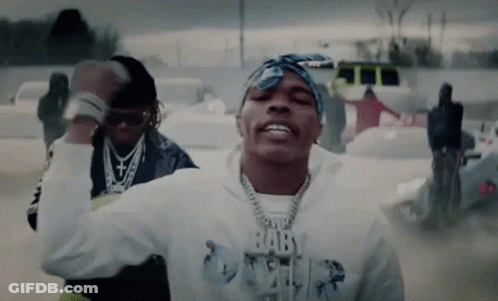 A Man Wearing A Bandana And A Hoodie With The Word Baby On It Is Dancing In A Parking Lot . GIF