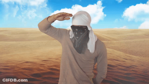 A Man Wearing A Bandana And A White Head Scarf Stands In The Desert GIF