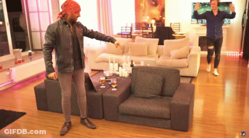 A Man Wearing A Bandana Stands In A Living Room Pointing At Something GIF
