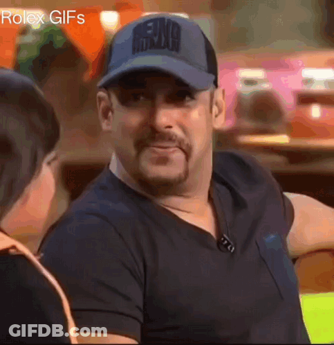 A Man Wearing A Baseball Cap And A Black Shirt Is Smiling While Talking To A Woman . GIF