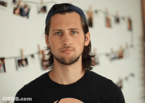 A Man Wearing A Baseball Cap And A Black Shirt Stands In Front Of A Wall With Pictures Hanging On It GIF