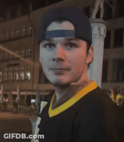 A Man Wearing A Baseball Cap And A Black Shirt With A Yellow Collar Looks At The Camera GIF
