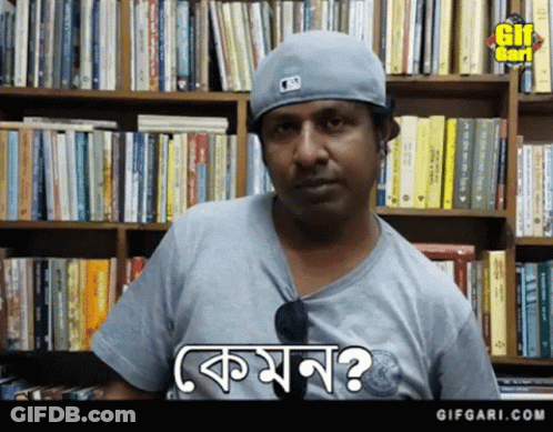 A Man Wearing A Baseball Cap And A Gray Shirt Is Standing In Front Of A Bookshelf And Says Komon GIF