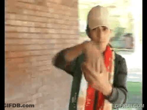 A Man Wearing A Baseball Cap And A Scarf Is Standing In Front Of A Brick Wall . GIF
