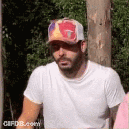 A Man Wearing A Baseball Cap And A White Shirt Is Standing In Front Of A Wooden Pole . GIF