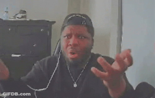 A Man Wearing A Baseball Cap Is Sitting At A Desk With His Hands Folded In Front Of His Face . GIF