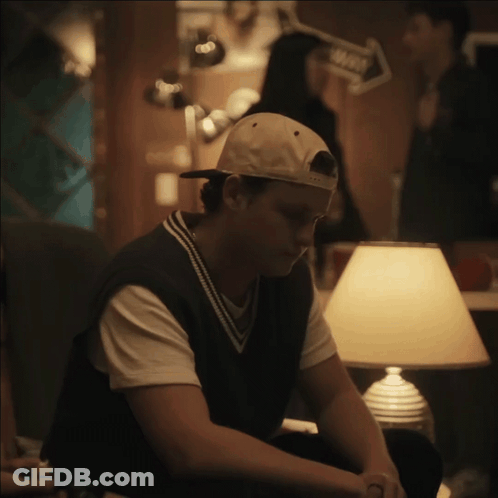 A Man Wearing A Baseball Cap Sits In Front Of A Lamp In A Dark Room GIF