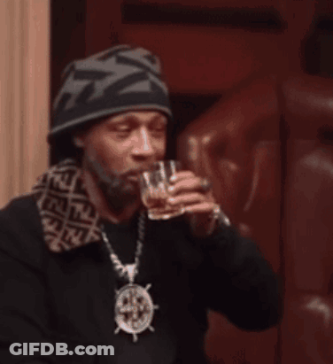 A Man Wearing A Beanie And A Necklace With A Steering Wheel On It Drinks From A Glass GIF