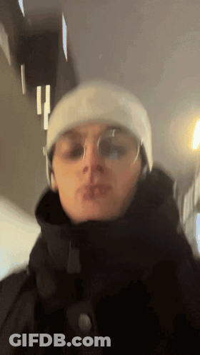 A Man Wearing A Beanie And Glasses Is Standing In A Dark Room GIF