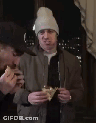 A Man Wearing A Beanie Is Eating A Sandwich While Another Man Watches GIF