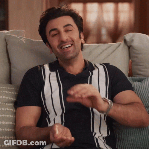 A Man Wearing A Black And White Striped Shirt Is Smiling While Sitting On A Couch GIF