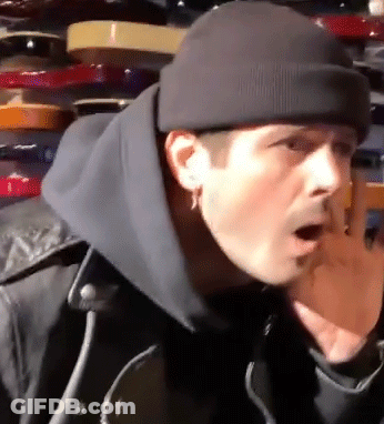 A Man Wearing A Black Beanie And A Black Jacket Is Standing In Front Of A Shelf . GIF