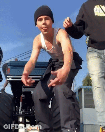 A Man Wearing A Black Beanie And A White Tank Top Stands Next To A Truck GIF