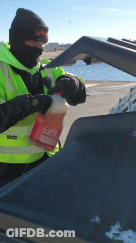A Man Wearing A Black Beanie And A Yellow Vest Holds A Gallon Of Orange Liquid In His Hand GIF